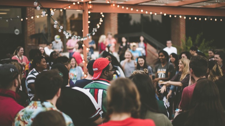 An outdoor gathering under string lights, people standing in groups and laughing.
